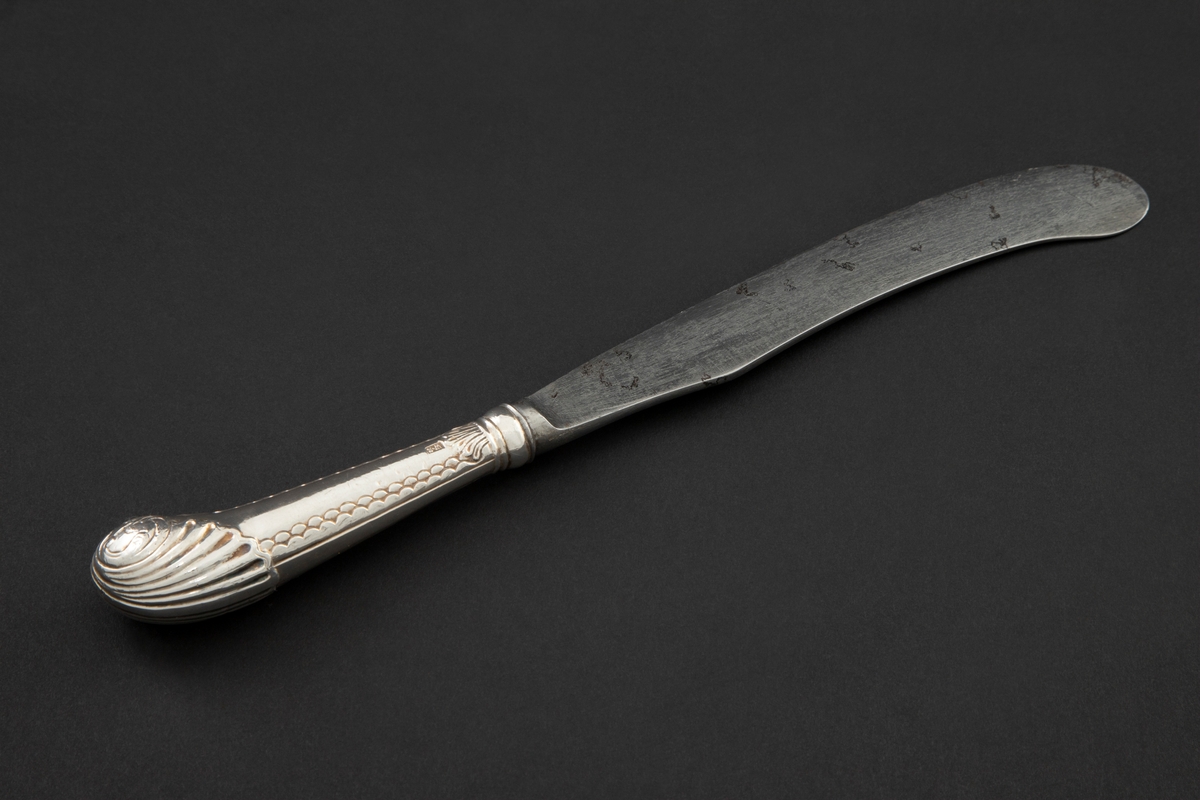 Kniv med sølvbelagt skaft og knivblad av stål. Knivbladet er flatt, buet med rund avslutning. Skaftet er rundt og dekorert med palmett- og bølgeornamentikk. Enden av skaftet er formet som et skjell med planteornamentikk.
