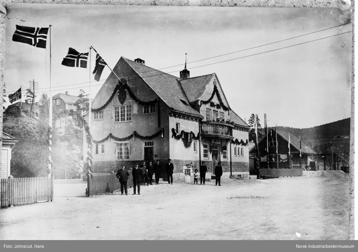 Gamle Notodden stasjon, sett fra framsiden. en gruppe mennesker står foran bygningen i uniform. Bygget er pyntet, det er heist flagg, og det henger et skilt med "1814-1914" over balkongen