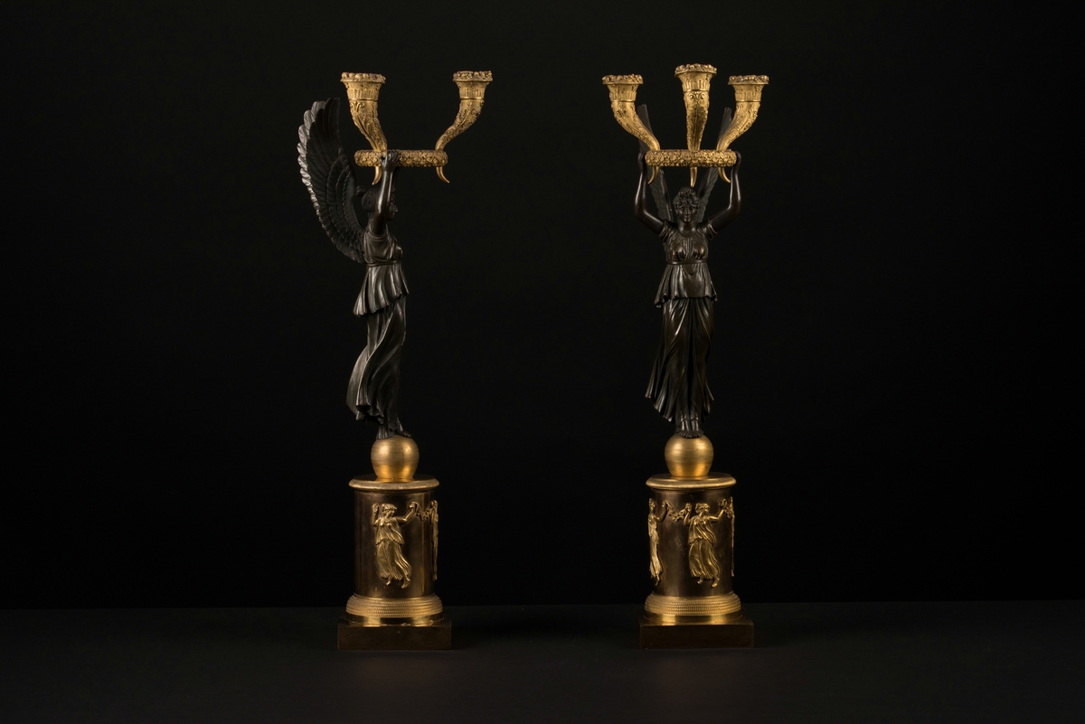Ett par brännförgyllda bronsljusstakar, kandelabrar, med tre ljushållare vardera.
En kolonnformad bas med ett klot på och en fyrkantig bottenplatta, av brännförgylld brons, bär upp en bevingad kvinna av svärtad brons. I de uppsträckta händerna sitter en krans med tre ymnighetshorn som utgör ljushållarna. Kransen och hållarna är även dessa av brännförgylld brons och dekorerade med akantusblad och vindruvsklasar. 
Runt den runda bärande basen finns reliefer som föreställer tre dansande kvinnor, de tre gracerna, som håller i blomstergirlanger/festonger.