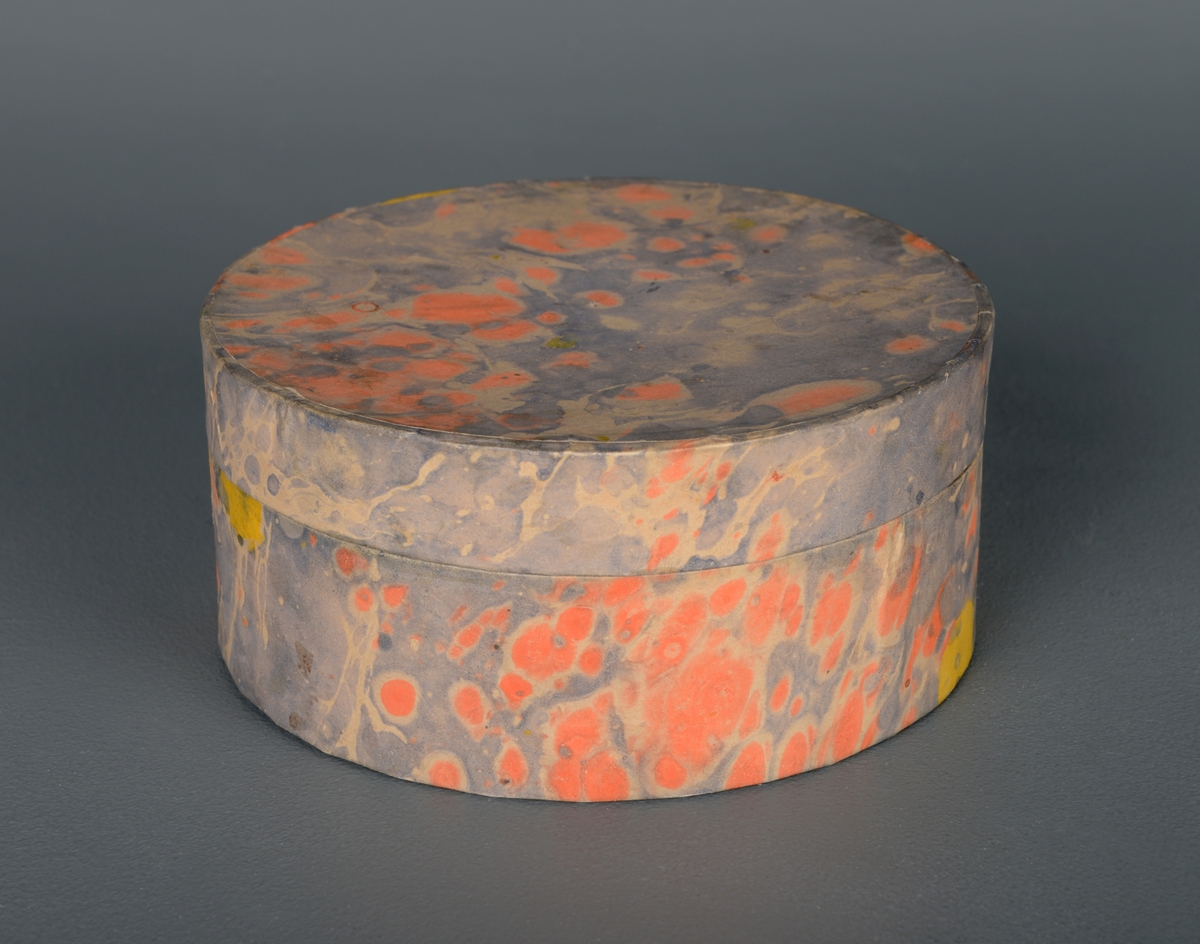 En rund, sylinderformet boks med lokk. Den er laget av papp, og trekt med papir. På utsiden er den grå, med gule og oransje prikker. I bunnen på innsiden og inne i lokket er den oransje med rødt marmorert mønster. På undersiden er den brun. Laget i pappsløyd.