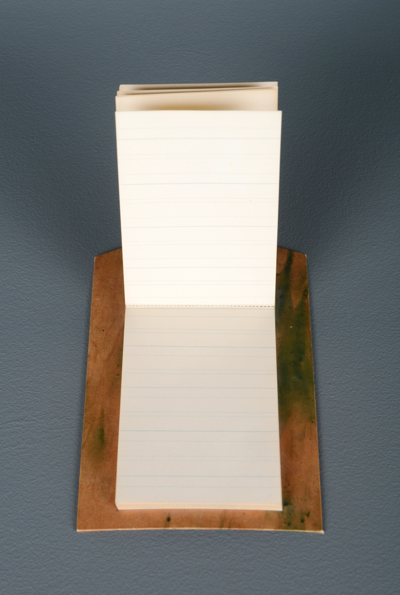 En liten, rektangulær notatblokk festet på en hard papplate. Papplaten er beige og grønn, og er avlang med buet topp. Det er et hull gjennom toppen av platen. På baksiden er det limt på papir. Laget i pappsløyd.