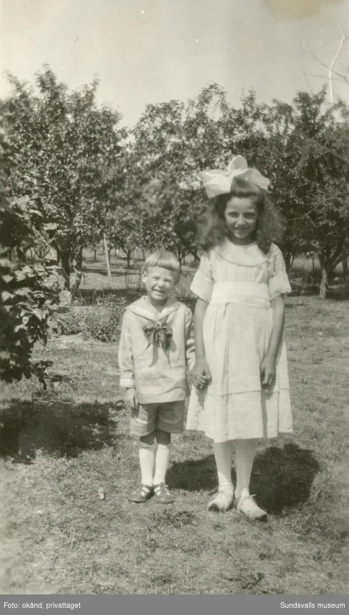 Två finklädda barn i en trädgård. På baksidan av bilden står det: Gunnar och Anna, (Waldemars flicka) 66. Ur ett album som tillhört Nils Hermansson, Svartvik.