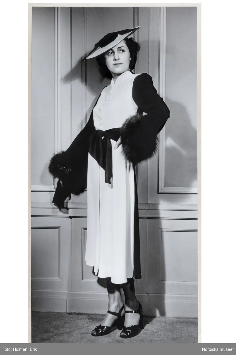 Mannekäng visar svartvit kappklänning med liten ståkrage, knäppning fram, knytskärp och ärm med pälskant. Liten hatt och remsandaler med vristrem. NK:s Franska damskrädderi våren 1938, möjligen från Jeanne Paquin.