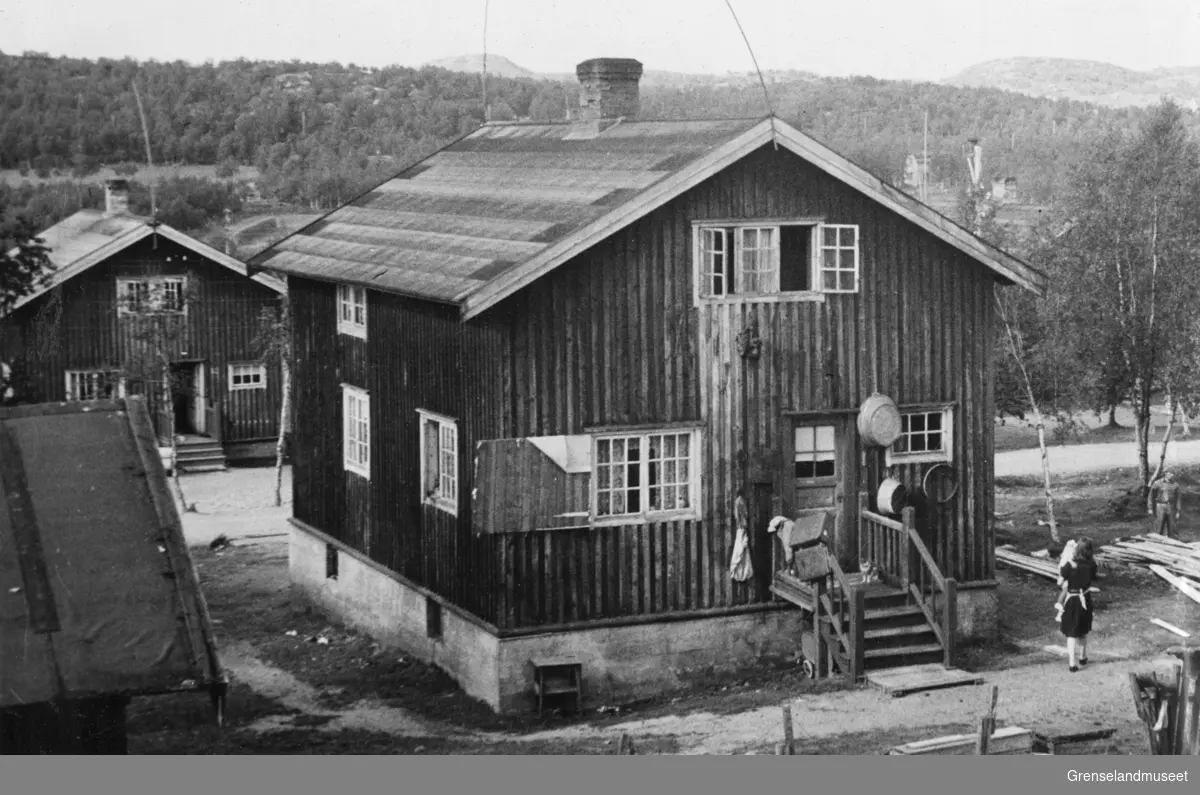 Gammelt egnehjem i Bjørnevatn, 19. august 1948. En kvinne med et barn på armen står og ser mot en mann foran en materialhaug.