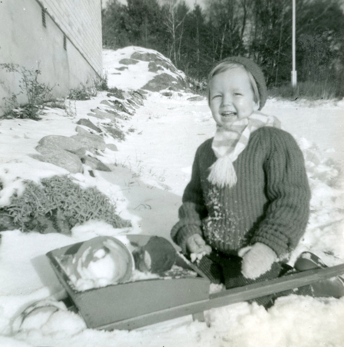Manne Ekman (född 1955) sitter och leker med sin skottkärra i snön utanför familjens bostad, Villa Maribo på Streteredsvägen 29 i Kållered år 1960.