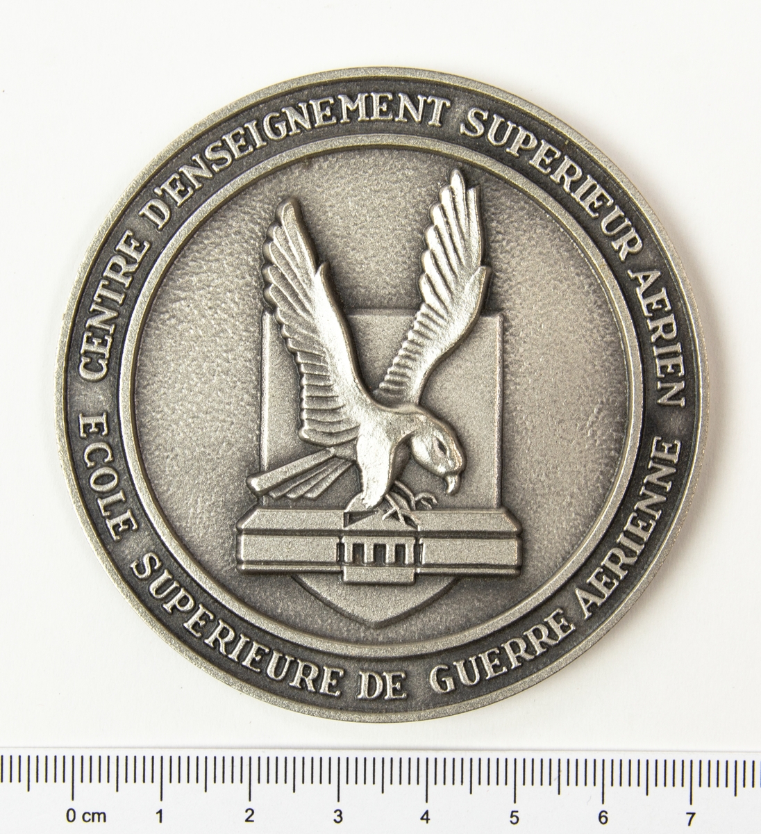 Rund medalj men en relief av en örn och ett hus och texten "Centre d'enseignement superieur aerien ecole supérieure de guerre aerienne" runt kanten. Medaljen förvaras i en ask.
