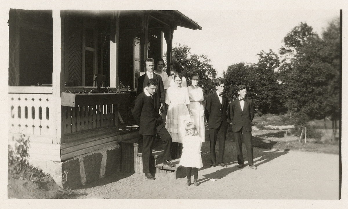 En grupp ungdomar och en liten flicka har samlats på trappan till en veranda. Ca 1920.