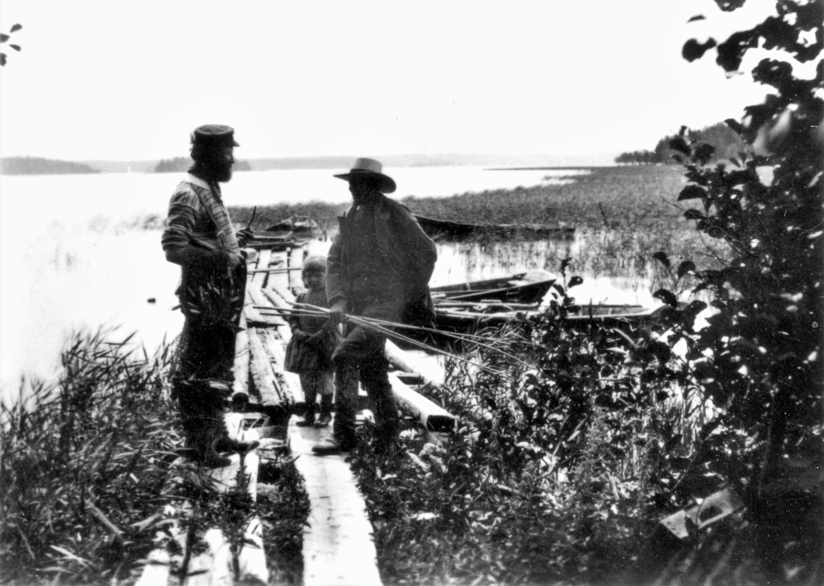 Två män och en flicka på en brygga, där ekor ligger förtöjda. Fiskare vid Knobesholmssjön. (Mannen till vänster finns även med på bild E5806)