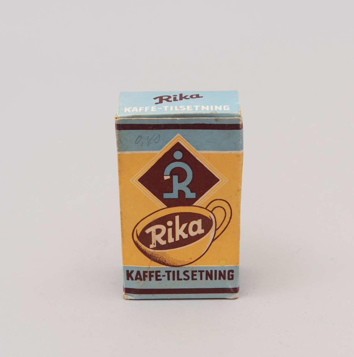 "Rika. Kaffe-tilsetning". Pappeske med innhold i matpapir. Påskrevet."0,40" (pris) med gråblyant. Produsert av A/S P.G. Rieber & Søn, Bergen. (motiv i gult, blått og brunt)