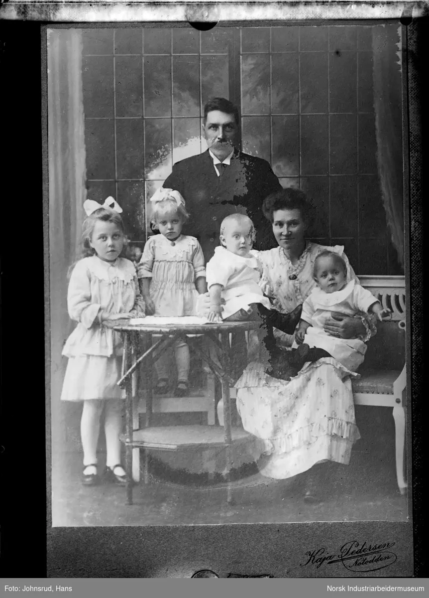 Avfotografert visittkort som viser en familie. En mann stående bak en sittende kvinne med to spedbarn på fanget og to stående jenter med sløyfer i håret.