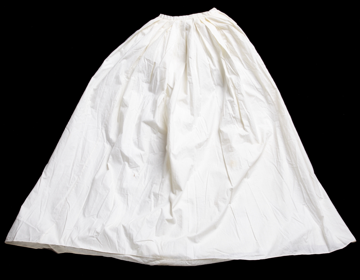Underkjol att bära under dopklänning M 66048:1, vit dopklänning. Tillverkad av fin bomullslärft med veckad, insydd midja med påsydd linning och långt sprund mitt bak. Knyts med de i linningen fastsydda bomullsbanden.
