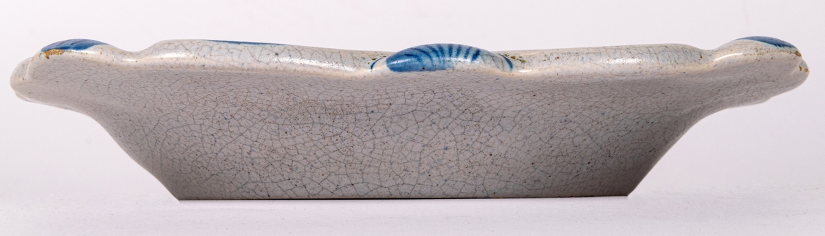 Fyrkantig assiett i lergods från Bo Fajans, modell 1084, formgiven av konstnär Allan Ebeling 1925. Glasyr AK: gråblåtonad antik konstglasyr (krackelerad) med handmålad dekor i blått i spegeln och på brämen.