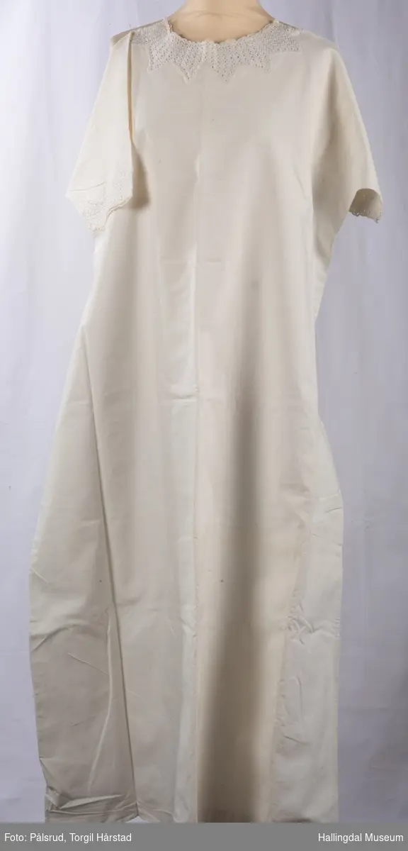 Hvit serk eller nattkjole i bomullsstoff med heklete kanter på hals om rundt ermene.