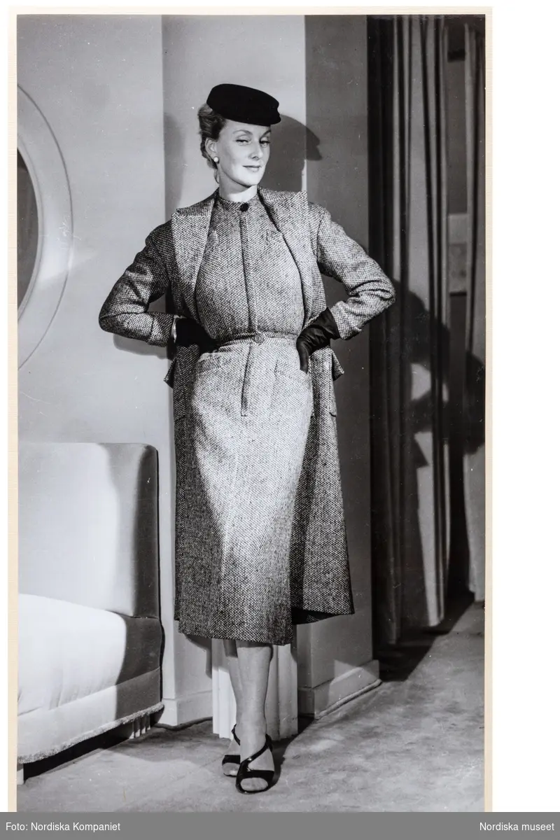 Mannekängen Margareta Berglund gift Nordlund (1946) visar höstplagg i provrummet vid NK:s Franska damskrädderi. Klänning eller dräkt av tweed med knäppning fram, markerad midja och snedfickor. Vadlång rak kjol. Över bär hon en kappa i samma tyg och längd. Hatt, handskar och remsandaletter.