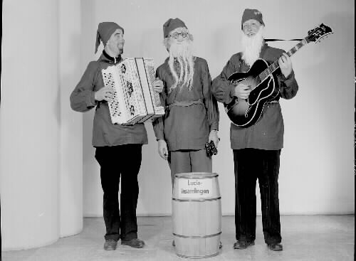 Tre musicerande tomtar; en med dragspel och en har gitarr. En tunna framför dem är märkt med "Luciainsamlingen". Beställare: Tage Sandberg.