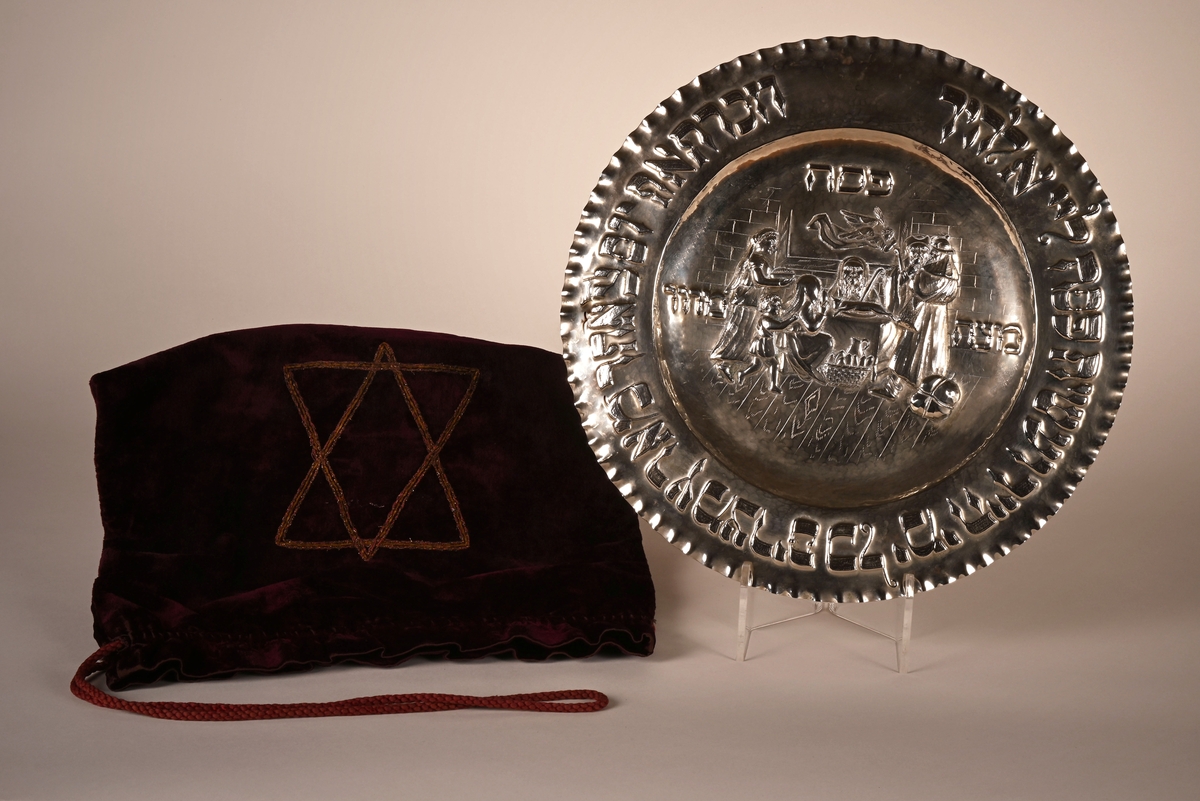 Sølvfarget fat med hebraisk skrift rundt kanten. I midten av fatet er det preget inn en ofringsscene: Et lam ligger på et bord, og fem personer er samlet rundt bordet.