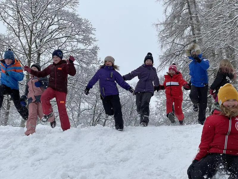 Ni barn løper og hopper i snøen i parken, store trær i bakgrunnen