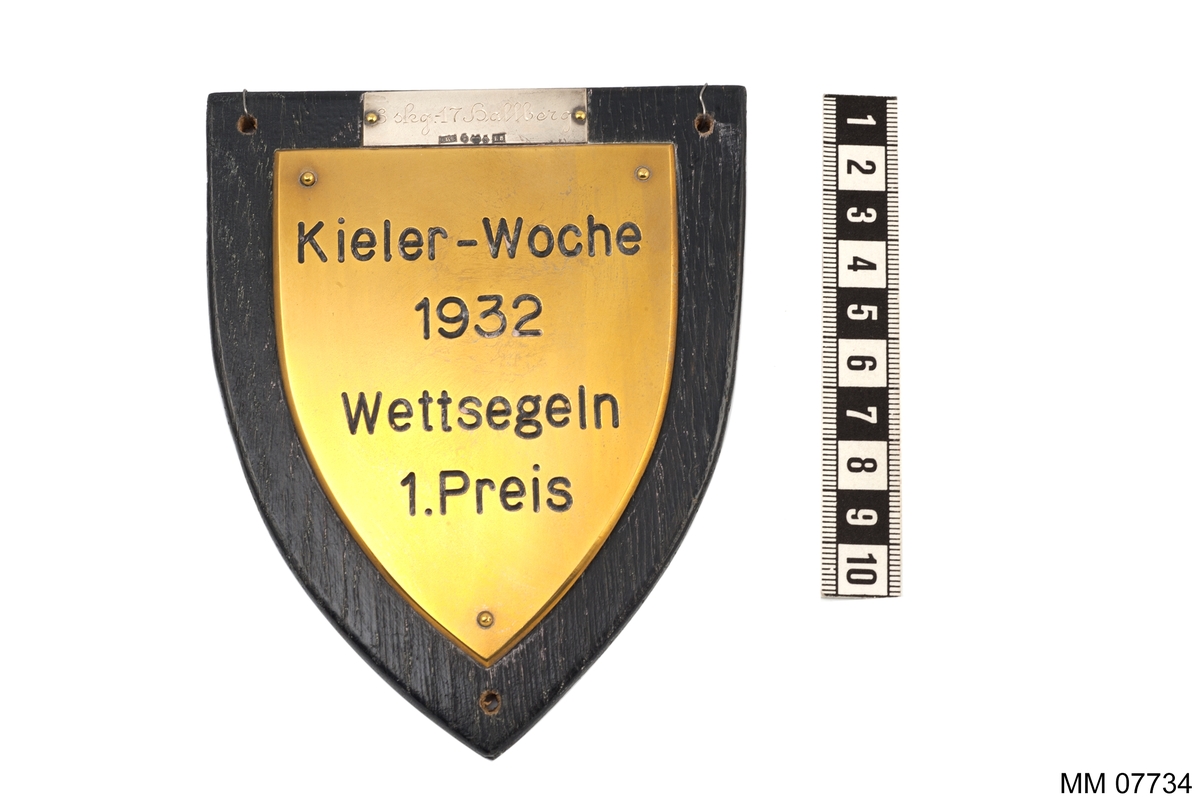 Plakett av brons på platta av trä, svart.
Inskription: Kieler-Woche 1932.
Wettsegeln I Preis.
På silverplatta: 3 skg,17 Hallberg.