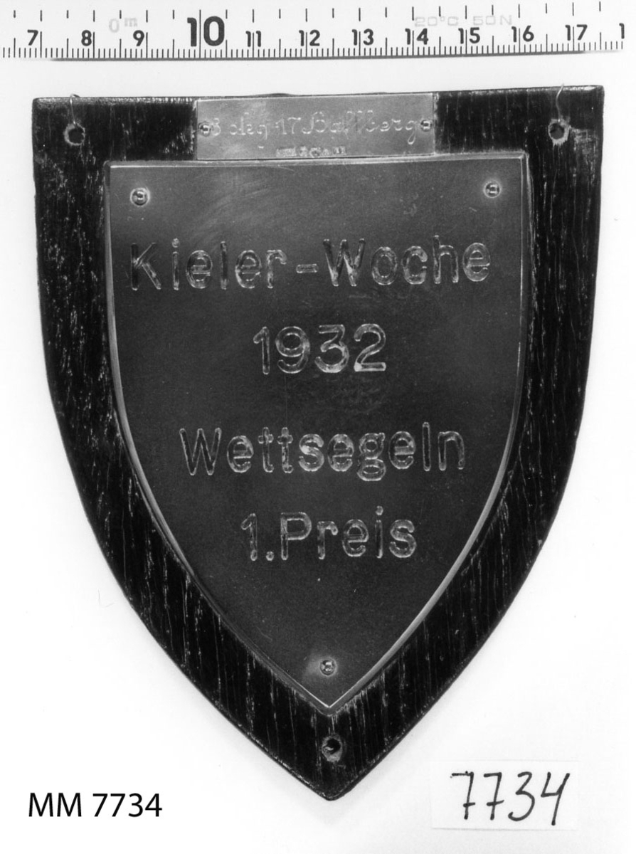 Plakett av brons på platta av trä, svart.
Inskription: Kieler-Woche 1932.
Wettsegeln I Preis.
På silverplatta: 3 skg,17 Hallberg.