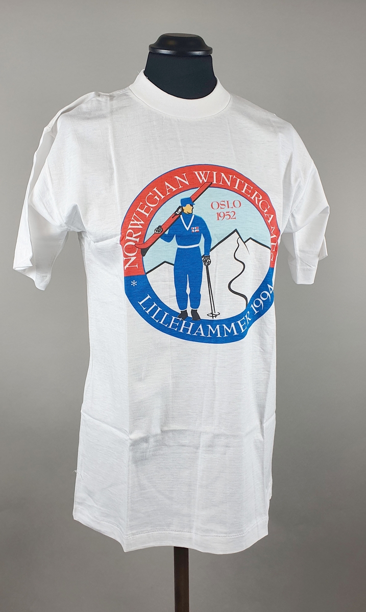 Hvit t-skjorte av bomull med et rundt bilde på brystet, med tegning av en skiløper med ski over skulderen og staver i hånden, kledd i klær fra 1950-tallet. Tekst rundt bildet.