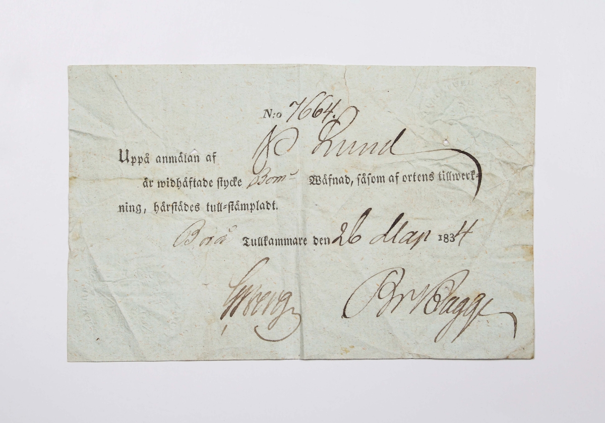 Tullsedel i gråblått papper. Liten reva upptill. Handskriven text i svart bläck: "N:o 7664 Uppå anmälan af P. Lund är vidhäftande stycke Bom-Wäfnad, såsom af ortens tillverkning, härstädes tull-stämpladt. Borås tullkammare den 26 dag 1834". Signerat av två namnteckningar. Försedd med tullverkets stämplar i Borås. Medföljer brev från givaren.