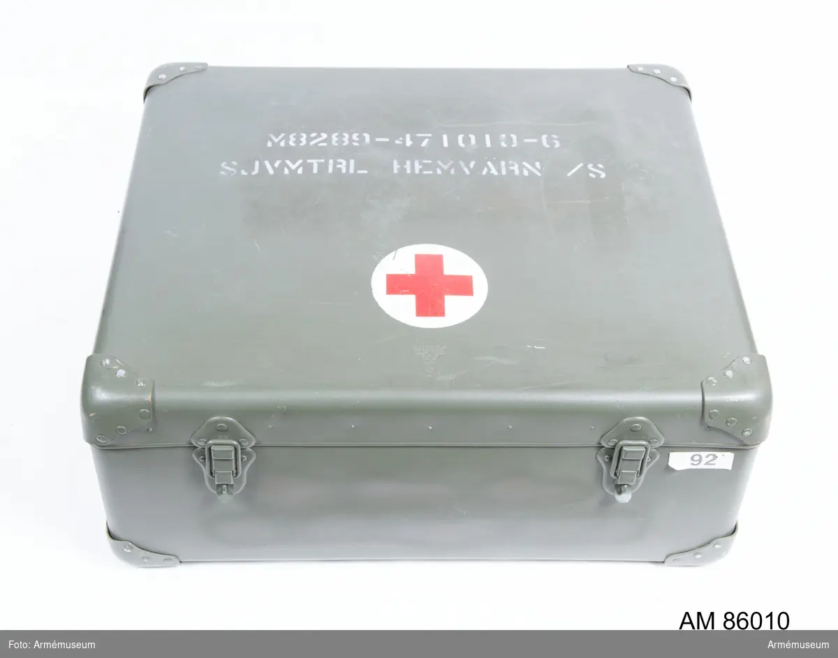 Sjukvårdslåda för Hemvärnet. På väskans ovansida står skrivet med vit text: 
"M8289-471010-6 SJVMTRL HEMVÄRN /S". Lådan är nästintill komplett och har följande oanvänt innehåll (se bifogad satslista för detaljer):
1 st innehållsförteckning (satslista)
1 st aluminiumask innehållande 2 st tvålar
1 st rulle innehållande 50 st avfallspåsar i plast á 30 liter
1 st Rödskorsflagga 100x75 cm
1 st peang Kocher 14 cm
1 st stickbäcken plast
1 st rondskål plast 25 cm
1 st förpackning plastbägare 20 cl för engångsbruk
1 st plastpåse fö förvaring av sjuktransportjournal
10 st sjuktransportjournaler (i påse)
1 st patientliggare
2 st handdukar
1 st nagelborste
5 st kirurgisk mikroporös tejp
10 st första förband
10 st binda elastisk 5 m x 8 cm
10 st binda elastisk 5 m x 12 cm
100 st tungspatlar (i ask)
2 st brännskadeförband 60x80 cm
1 st vattentätt tyg
2 st febertermometrar
10 st nummerlappar utr död
30 st snabbförband 80x20-25
1 st snabbförband elastisk 5 m x 6-8 cm
1 st rulle hushållspapper
1 st kittel aluminium med lock
1 st förbandssax trubbig
6 st självhäftande förband