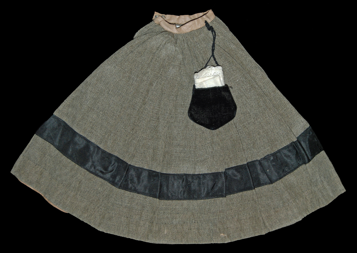 Kjol, grågrön med svart band påsytt på nedre delen av kjolen. Beigt band i midjan. Svart sammetsväska med band fastsydd i linningen på kjolen, innehållande en liten spetsnäsduk.