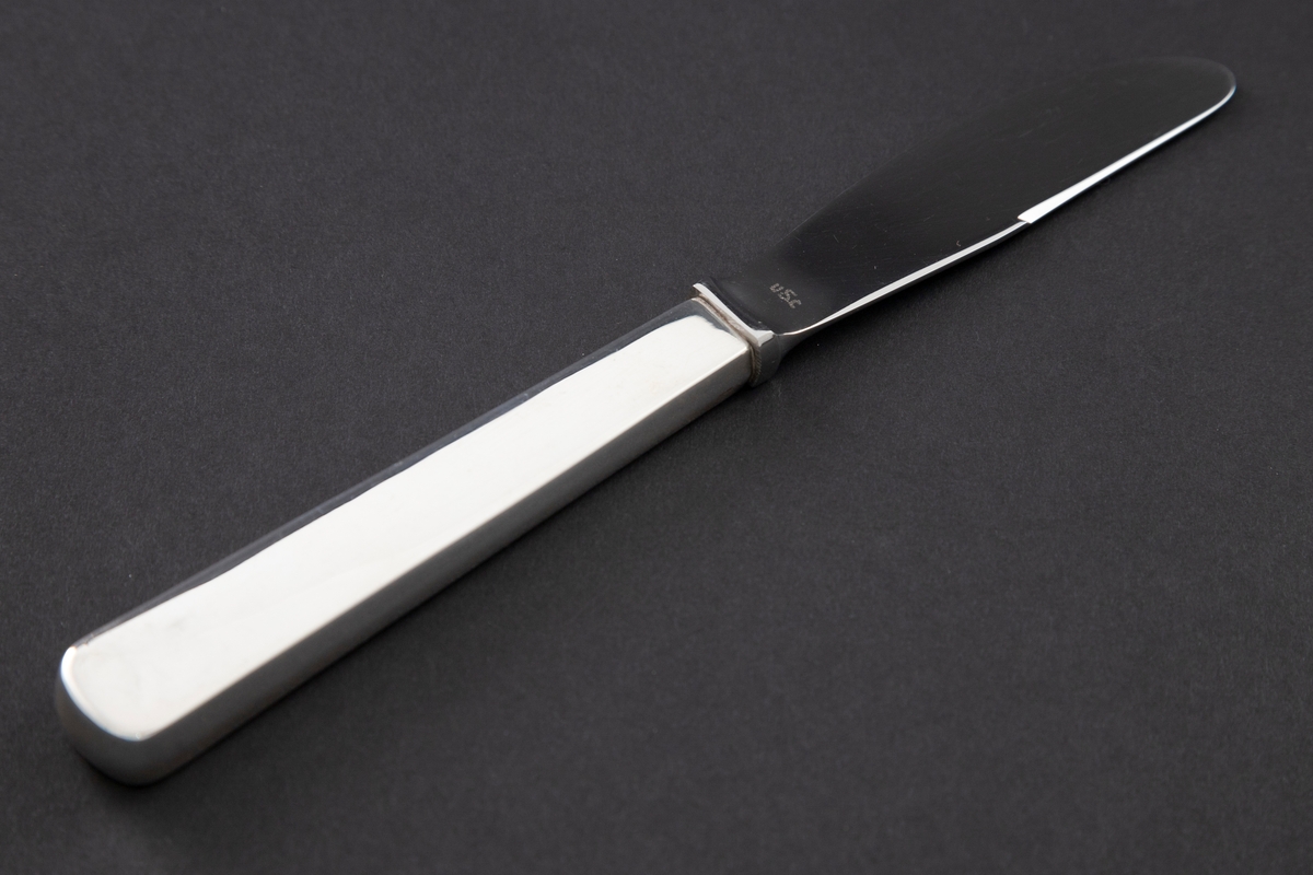 Dessertkniv med skaft i sølv. En rektangulær holk markerer overgangen mellom det lange, tykke skaftet og bladet. Sistnevnte er utført i rustfritt stål. Glatt udekorert overflate.