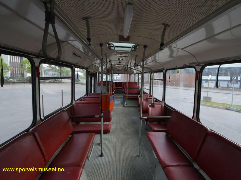 Buss med rødt eksteriør. Det er to inngangs- og utgangspartier på bussen. Bussens interiør består av røde enkelt og dobbeltseter og lyse overflater på tak og gulv.