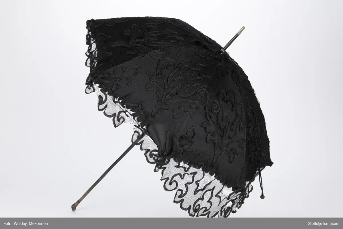 Parasoll med 8 spiler av metall og treskaft. Parasoll er trukket med svart stoff og en utenpå liggende svart tyll med dekor.