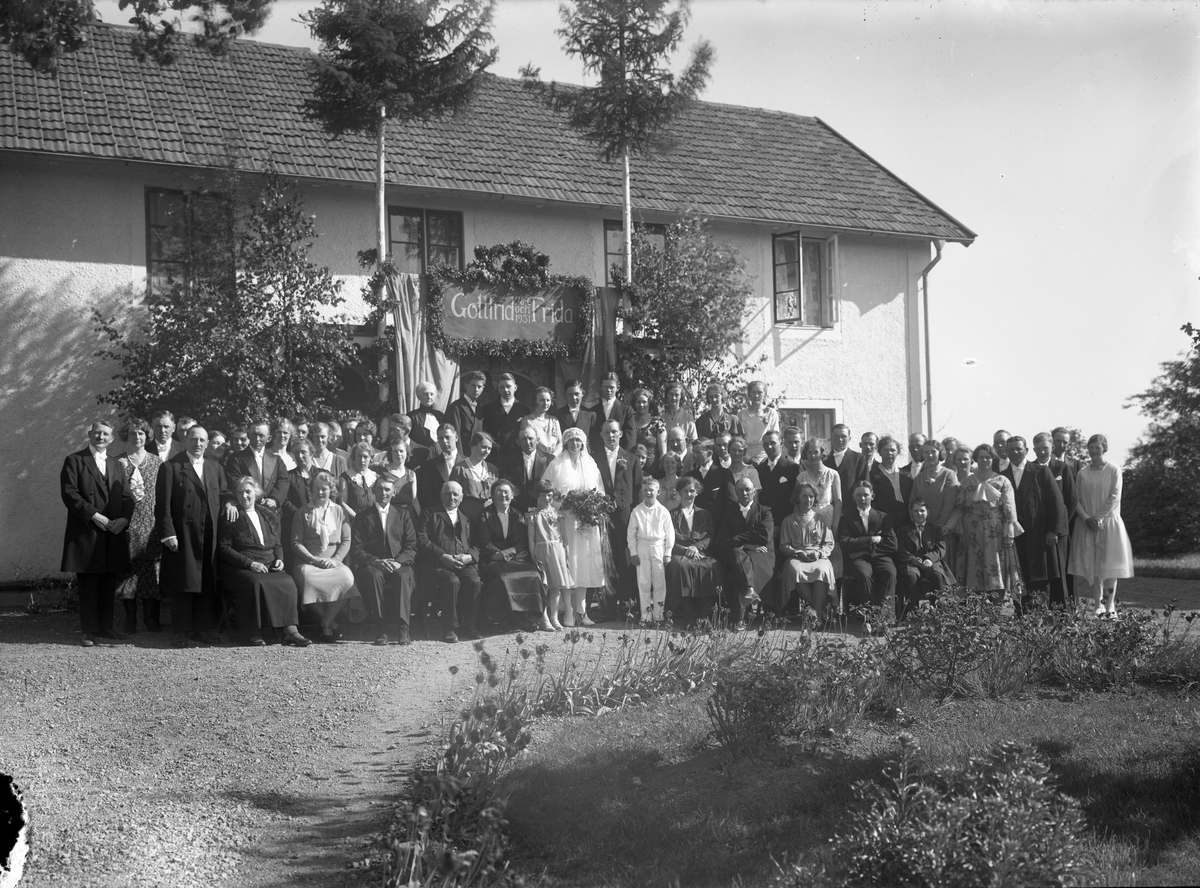 En stor grupp kvinnor, män och barn uppställda framför en ljus pustad envåningsbyggnad. I centrum står brudparet Gottfrid och Frida under en lövad skylt med deras namn och datumet 26/6 1931.