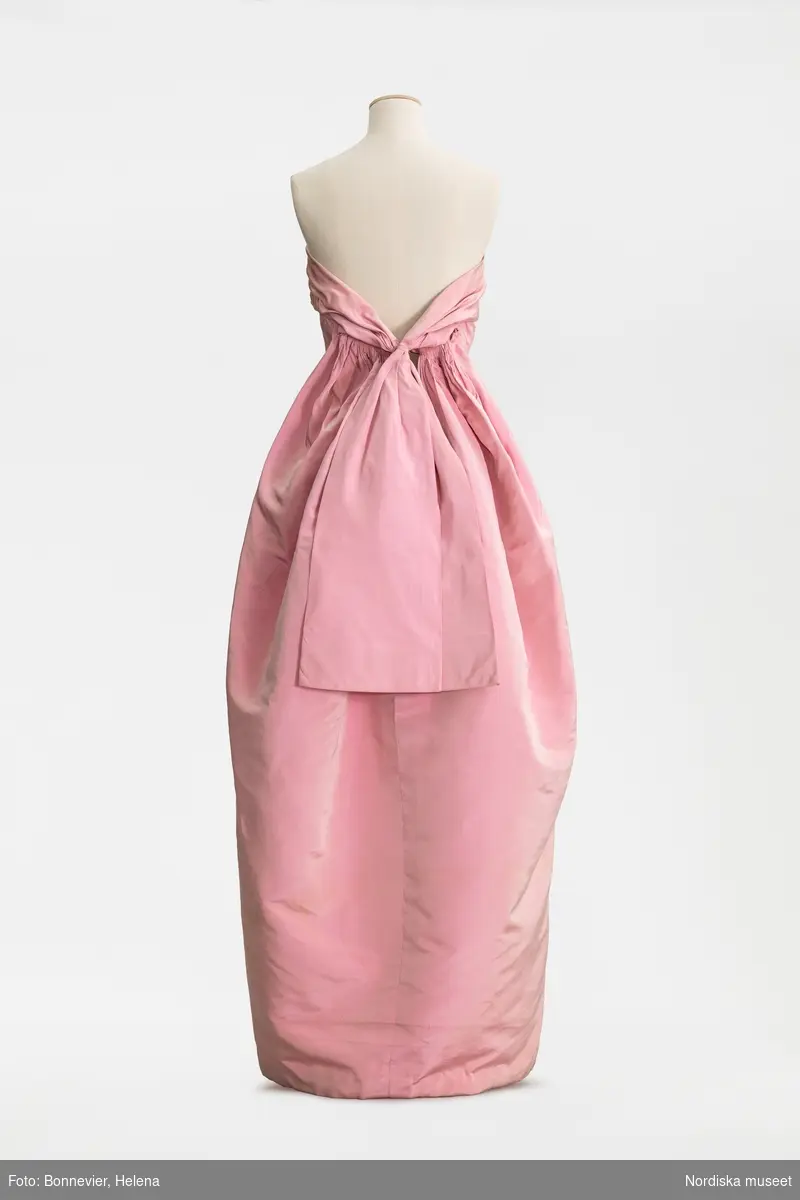 Klänning monterad på docka. Originalmodell från Cristóbal Balenciaga i rosa sidenfaille som visades på NK:s Franska damskrädderi. Karin Ellhammar (född 1938) erbjöds att köpa originalet i slutet av säsongen, hösten 1960. I Röhsska museets samlingar, RKM 40-1999.