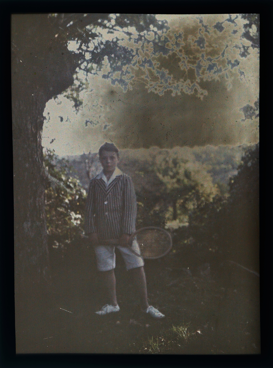 Portrett av en gutt med en tennisracket i hånden, ved siden av et tre og vegetasjon.