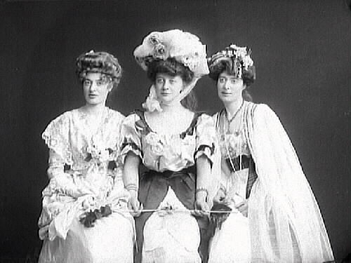 Tre kvinnor i exklusiv klädsel, möjligen utländskor. Kanske är de aktriser. Hon i mitten bär en enorm hatt, urringad, kortärmad klänning och håller i en dirigentpinne eller liknande. 
(Se även MR2_929, MR2_969 och MR2_931, MR2_975, MR2_976)