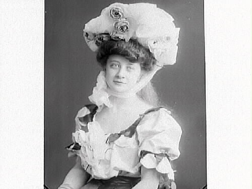 Ateljéporträtt av en ung kvinna i en oerhört stor hatt fäst med band under hakan och en matchande kortärmad klänning med puffärmar och decolletage. Kanske en aktris av något slag? (Se även bilderna MR2_931, 950, 969, 975)