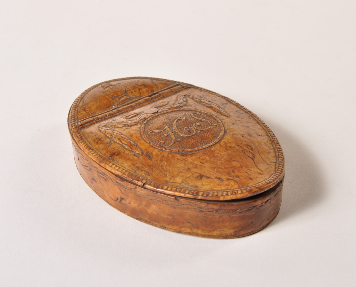 Oval boks av valbjørk, med hengslet lokk. Utskåret mønster på lokk: urne girlander og monogram.