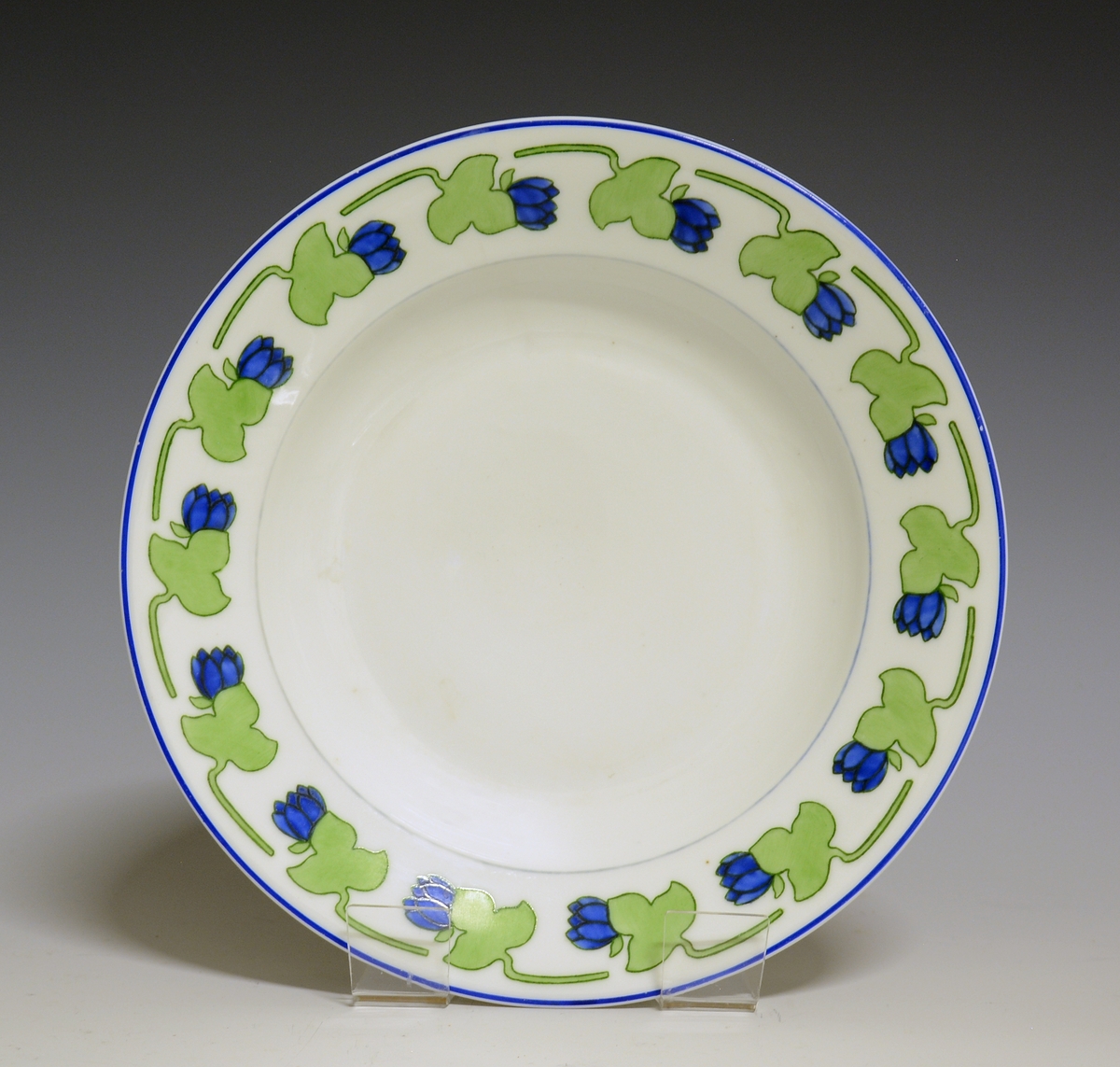 Dyp tallerken i porselen. Hvit glasur. Stilisert bord med blå blomst og grønt blad rundt  fanen.
Modellnr: 15.2