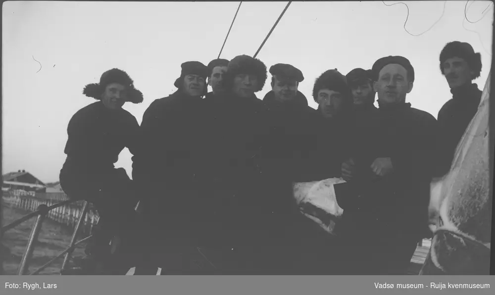 Et gruppebilde av ni sjømenn, på dekket til en båt. Båten ligger ved en trekai. Til venstre i bakgrunnen, ses noen bygninger. Flere av mennene har skinnlue  på hodet. På seilet har det lagt seg litt snø. Bildet har tilhørt Lars Rygh fra Hønefoss, som var med på minerydding i Varangerfjorden, mellom 1917 - 1918.