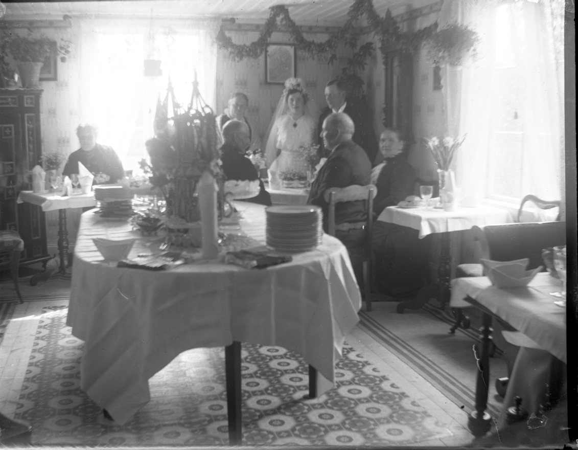 Hemmiljö med ett brudpar stående längst bort i rummet under en girlang bakom ett bord med vit duk. I framkant står ett bord med vit duk och staplade tallrikar bland annat. Flera små bord med vita dukar. Två män och tre kvinnor står resp sitter i brudparets närhet.