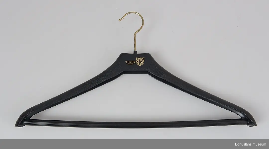 Klädgalge för byxa av svart plast med påtryck i guldfärg av firmamärke TIGER OF SWEDEN med företagssymbol.