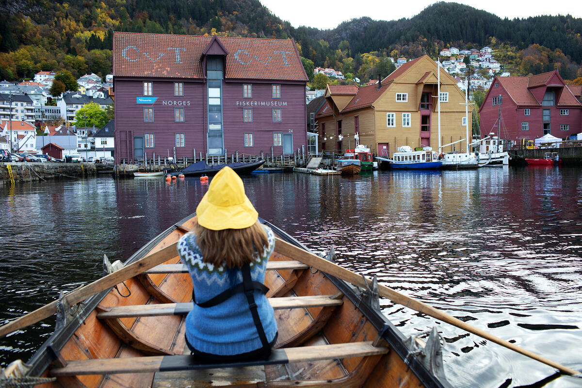 Vi ser en dame som sitter med redningsvest og gul sydvest og ryggen til oss, mens hun ror en tradisjonsrobåt. Foran henne er Norges Fiskerimuseum