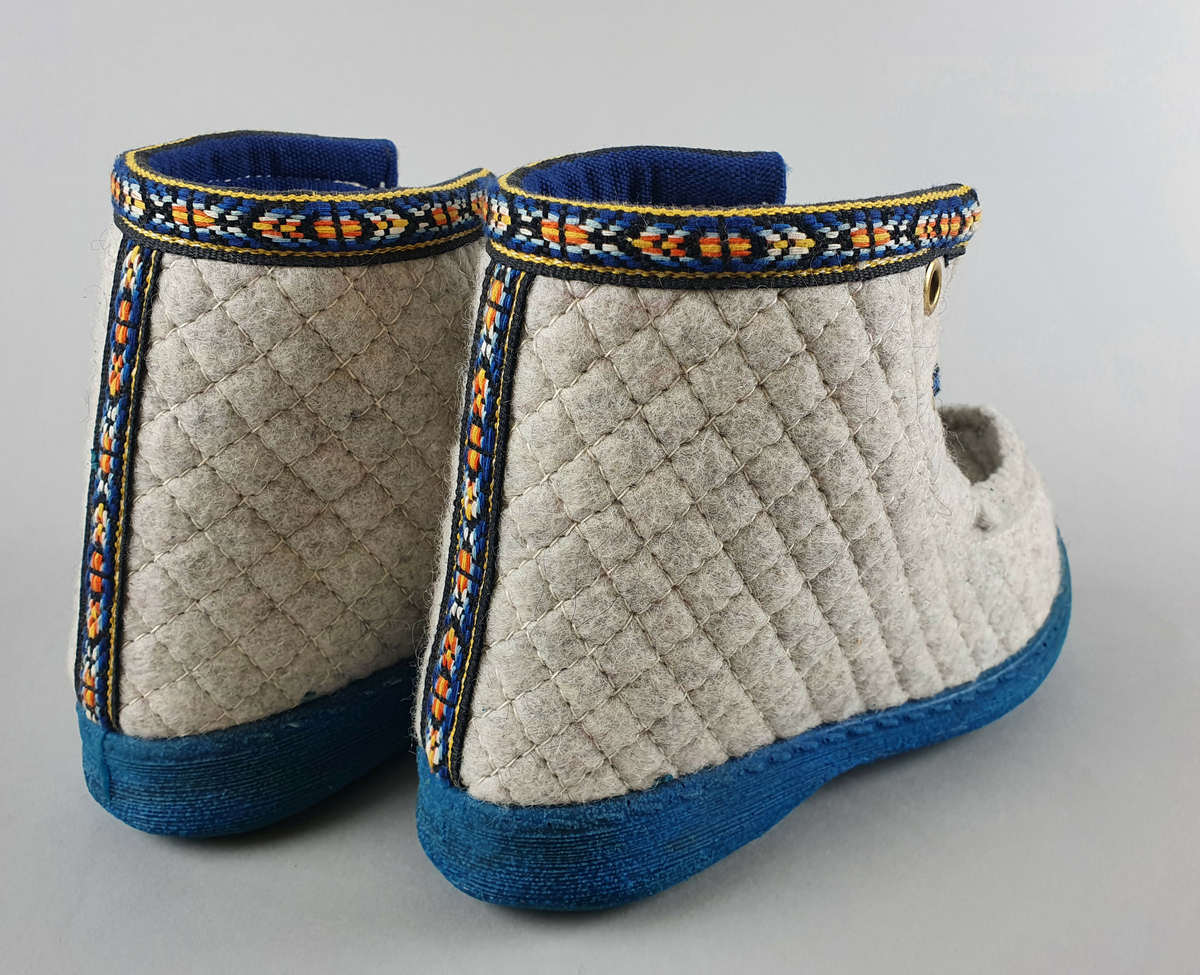 Barnestøvler (Nesnalobben) for vinterbruk av ullfilt med blå såle av gummi, blå lisser og mønstret bånd rundt kanten øverst og midt bak. Den ene skoen mangler et lissehull.