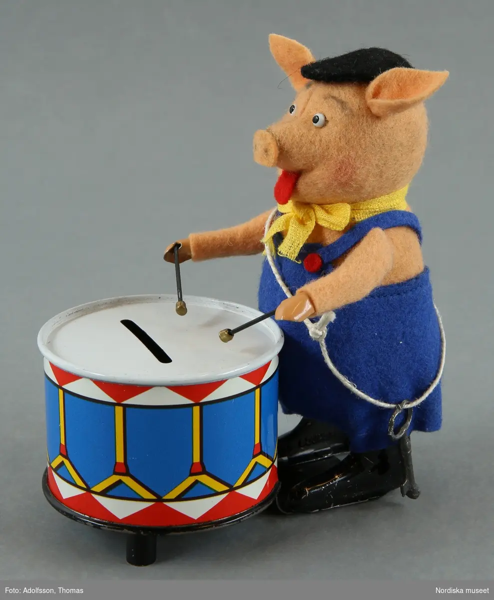 Inventering Sesam 1996-1999:
Gris  H  12       Ask  L  11,3   B  6,5   H  12 (cm)
Gris spelande på trumma (sparbössa), fjäderdriven leksak, av plåt, grisen klädd med filt, blå hängselbyxor, gul rosett om halsen och svart keps, svartlackerade skor, trumman litograferad i vitt, blått rött och gult, nyckel i snöre. Märkt: SCHUCO PATENT/ MADE IN GERMANY".  Grisen lyfter armarna och spelar på trumman, nyckel till fjäderverket saknas.
Gul originalask av papp med text och bild i flerfärgstryck, motiv av gris från Walt Disney's film "Tre små grisar",  text bl a: "WALT DISNEY / 980/5 / Schuco / Made in Western Germany". Påklistrad prislapp: "8.75". 
Walt Disney-grisar från Schuco, se Battenberg Antiquitäten-Katalog Schuco, av Rudger Huber, Tyskland, 1995, s 140-141.
Anna Womack jan 1998