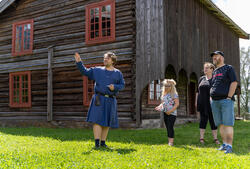Guide i middelalderdrakt står foran et laftet, gammelt hus og peker og forteller til en familie. (Foto/Photo)