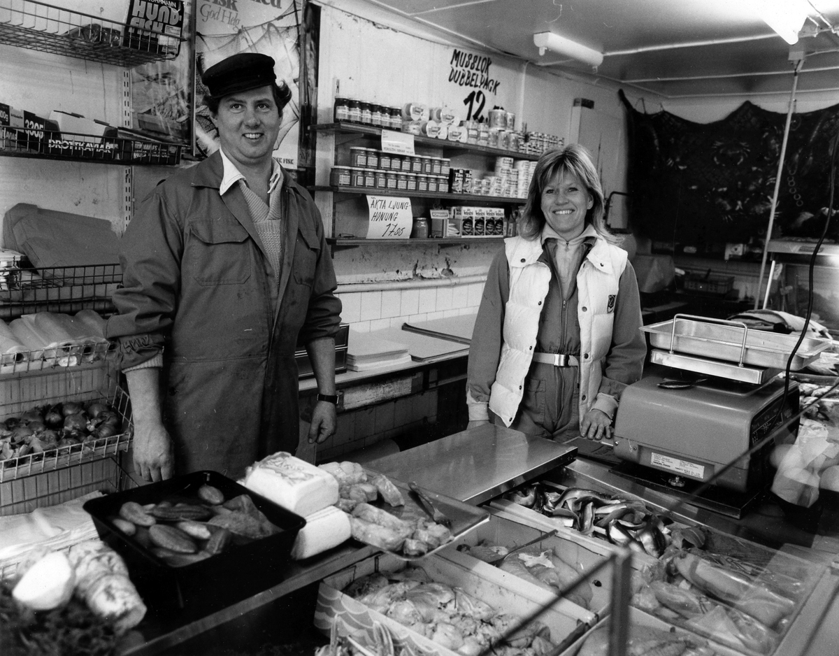 En man och en kvinna bakom disken på Västkustens fiskaffär. På väggen bakom ses reklam för bland annat musslor och honung. Fastigheten revs i månadsskiftet juli-augusti 1984.