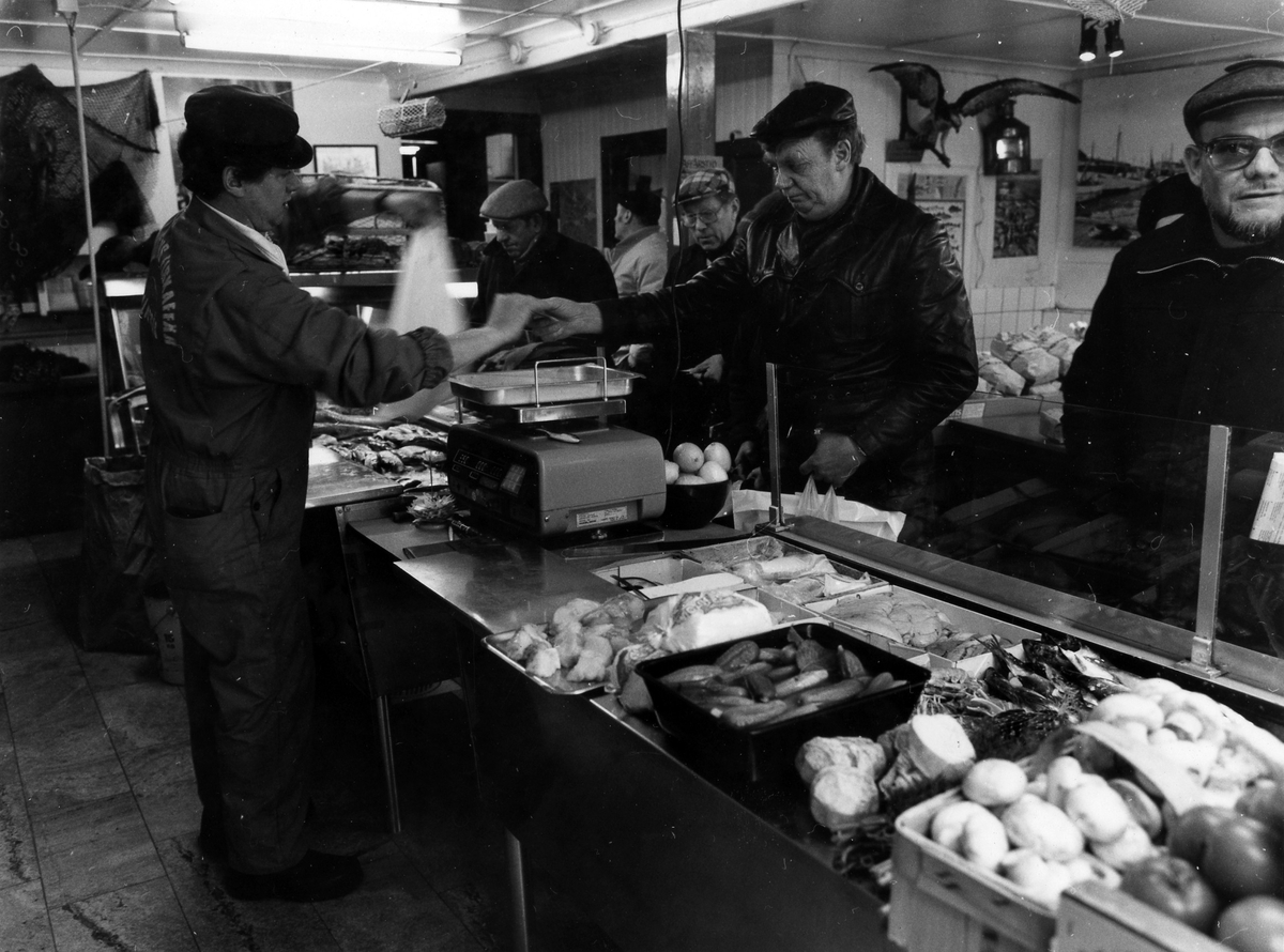 På Västkustens fiskaffär ses kunder betjänas av en man bakom disken. Bakom glasrutan på disken ses ett sortiment av grönsaker, fisk och skaldjur. Fastigheten revs i månadsskiftet juli-augusti 1984.