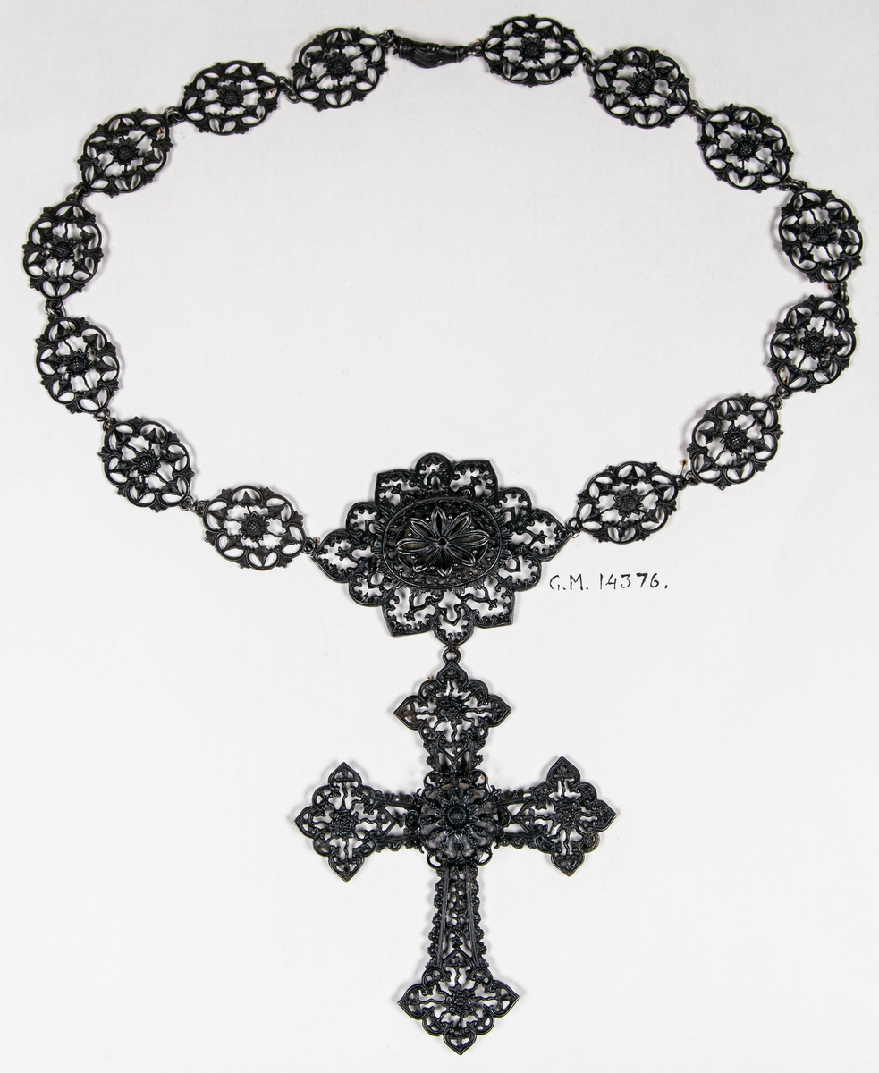 Smycke, halsband av metall, gjutet, genombrutna plattor, i mitten en större varvid ett kors är fastsatt. Lås: två händer som fattar varandra.