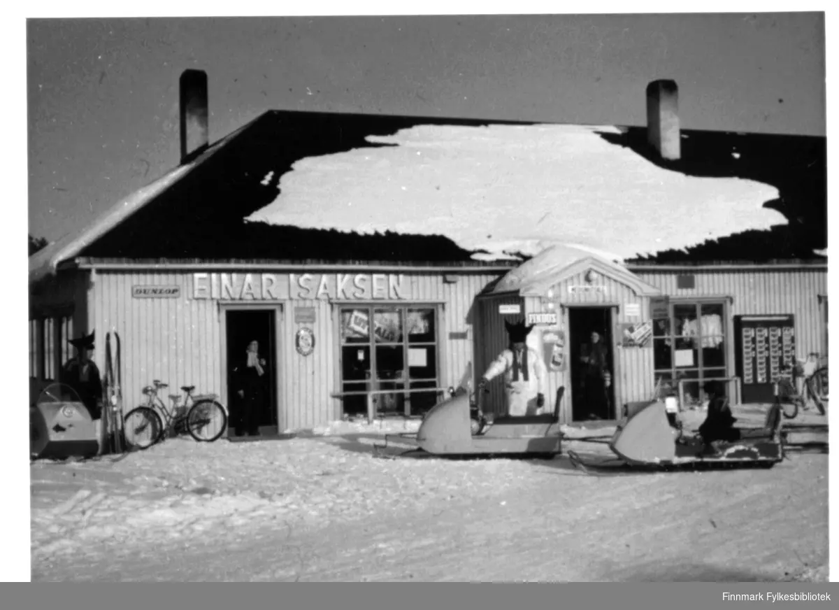 Tre snøscooter utenfor Einar Isaksen sin butikk i Karasjok, fotografert slutten av 1950-tallet/tidlig 1960-tallet. I hjørne med varen, antakelig Lars Larsen Gaup. Sjokoladeautomat på veggen. Dunlop dekk ble det også solgt, vi ser skiltet på veggen.