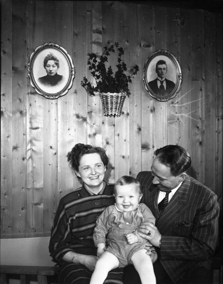 Kvinne i stripete kjole og mann i dress med et barn på fanget. To portrettbilder og en plante henger på veggen over dem. 