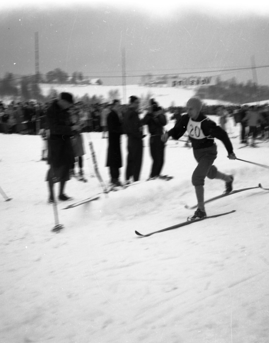 En skiløper med startnummer tyve i full fart i løypa. Mange tilskuere sees i bakgrunnen.